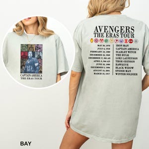 May include: A light green t-shirt featuring a graphic of Captain America with the text "CAPTAIN AMERICA THE ERAS TOUR" on the front. The back of the shirt has the text "AVENGERS THE ERAS TOUR" with a list of dates and characters.
