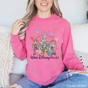 May include: A bright pink long-sleeve sweatshirt featuring a graphic of Disney's Toy Story characters and a castle. The words "Walt Disney World" are printed in a cursive font below the graphic. The sweatshirt is made of a soft material.