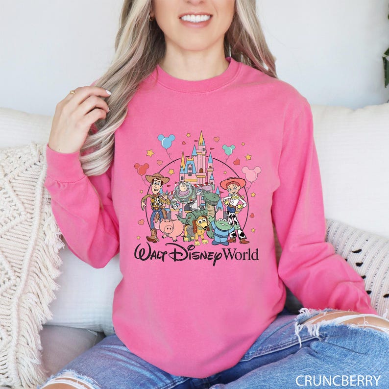 May include: A bright pink long-sleeve sweatshirt featuring a graphic of Disney's Toy Story characters and a castle. The words "Walt Disney World" are printed in a cursive font below the graphic. The sweatshirt is made of a soft material.