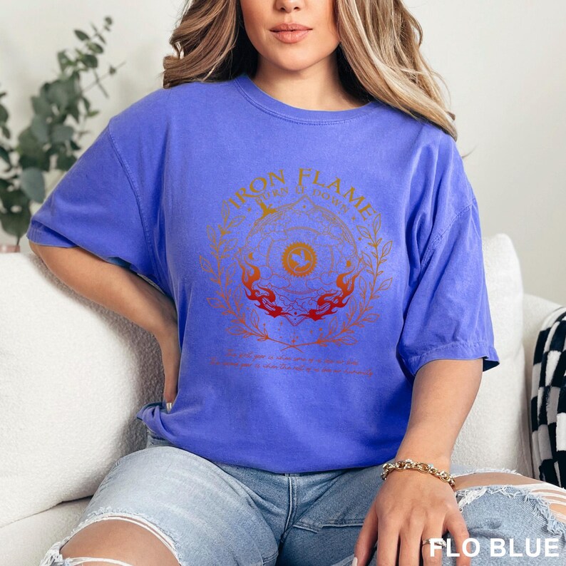 May include: A periwinkle blue short-sleeve t-shirt featuring a circular graphic in orange and red tones. The design includes the text Iron Flame, a central dragon emblem, and a surrounding wreath of leaves with flames at the base.