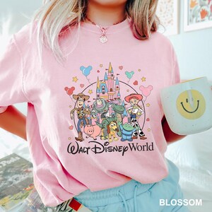 May include: A light pink t-shirt featuring a graphic of Disney's Toy Story characters and a castle, with the text "Walt Disney World". The shirt also has a white mug with a yellow smiley face.