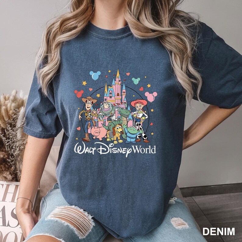 May include: Denim blue t-shirt featuring a colorful graphic of Disney characters, including Woody, Buzz Lightyear, and others, in front of a castle. The words "Walt Disney World" are printed below the graphic.