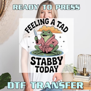 May include: White t-shirt featuring a cartoon frog wearing a pink robe and holding a knife. The text on the shirt reads "Feeling a Tad Stabby Today". The words "Ready to Press" and "DTF Transfer" are also visible.