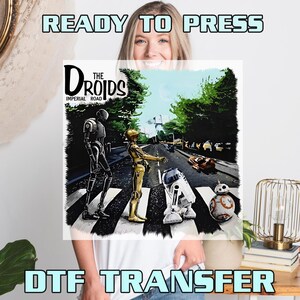 May include: DTF transfer featuring a parody of the Abbey Road album cover with Star Wars droids. The image includes the text "READY TO PRESS" and "DTF TRANSFER". The droids are crossing a street with trees and a vehicle.