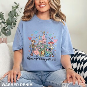May include: Light blue t-shirt featuring a colorful graphic of Disney characters and a castle, with the text "Walt Disney World". The shirt has a crew neck and short sleeves. The bottom of the shirt reads "Washed Denim".