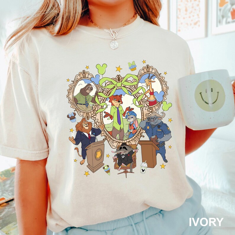 May include: Ivory-colored t-shirt featuring a colorful cartoon graphic of animal characters in a framed design. The graphic includes a sloth, fox, lion, rabbit, and other characters. The shirt also has a coffee mug with a smiley face.