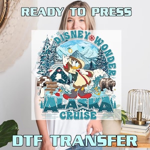 May include: White t-shirt featuring a colorful graphic of Donald Duck in winter attire, with the text "Disney Wonder Alaska Cruise." The design includes snowy mountains, a bear, and an igloo. The words "Ready to Press" and "DTF Transfer" are also visible.