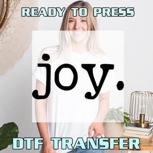 May include: White t-shirt with the word "joy" in large, black letters. The text "READY TO PRESS" and "DTF TRANSFER" are above and below the main text in a light blue font. The shirt is being worn by a person.