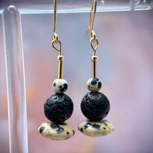 May include: Gold-tone dangle earrings with a layered design. Each earring features a small, speckled bead, a black, textured sphere, and a speckled, flattened oval. The earrings are suspended from gold-colored hooks.