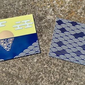 May include: Two rectangular coasters. One features a yellow sky with white clouds and a blue ocean with a sun graphic. The other coaster has a repeating blue and white wave pattern. Both coasters have a black border.