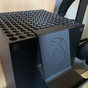 May include: Black Xbox Series X console with a matching black headset stand. The stand features the Xbox logo. The console has a perforated top for ventilation. The headset is black.