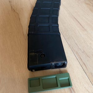 May include: A black, curved magazine with a textured surface and a separate olive green base plate. The magazine is rectangular with a series of raised rectangular patterns. The base plate has three recessed sections.