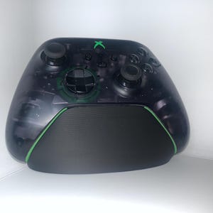 May include: A black and transparent Xbox controller with green detailing. The controller features a black base and green edging. The Xbox logo is green. It has two joysticks, a directional pad, and various buttons.