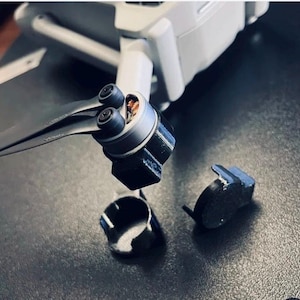 Op de afbeelding: Close-up van een drone met een zwarte propeller en motorbescherming. De arm van de drone is uitgeschoven en de propeller is bevestigd. Twee zwarte motorbeschermers en een zwarte propellerbeschermer liggen los op een donkere ondergrond.