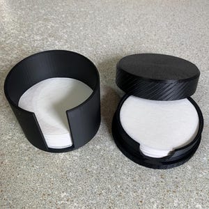 Può includere: Due supporti neri, stampati in 3D, per filtri di carta rotondi. Uno è aperto, cilindrico e contiene una pila di filtri bianchi. L'altro è un contenitore con coperchio e una pila di filtri bianchi all'interno.