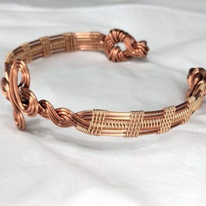 May include: A copper wire cuff bracelet featuring intricate woven patterns, twisted wire sections, and decorative coiled knots at both open ends. The metal has a polished, warm metallic finish with detailed craftsmanship throughout the design.