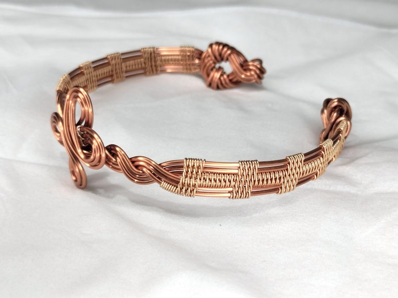 May include: A copper wire cuff bracelet featuring intricate woven patterns, twisted wire sections, and decorative coiled knots at both open ends. The metal has a polished, warm metallic finish with detailed craftsmanship throughout the design.