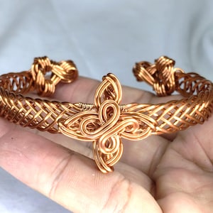 May include: A copper wire cuff bracelet featuring a woven lattice pattern along the band and a central decorative element shaped like a four-petalled flower or knot. The ends of the cuff are finished with coiled wire loops.