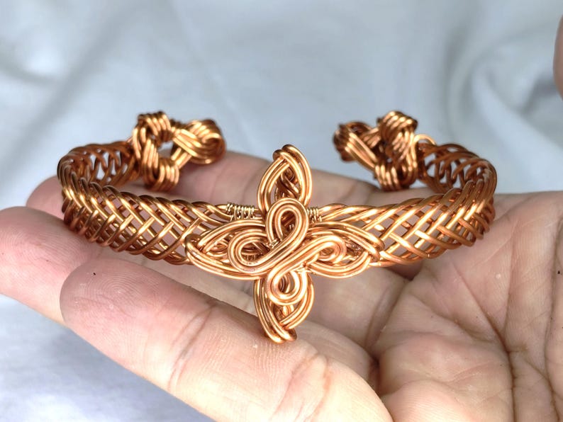 May include: A copper wire cuff bracelet featuring a woven lattice pattern along the band and a central decorative element shaped like a four-petalled flower or knot. The ends of the cuff are finished with coiled wire loops.