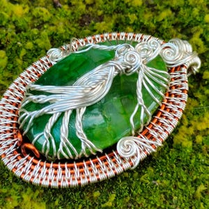 May include: A green gemstone pendant with silver wire wrapped around it, set in a copper wire frame. The pendant is oval shaped and has a smooth, polished surface.