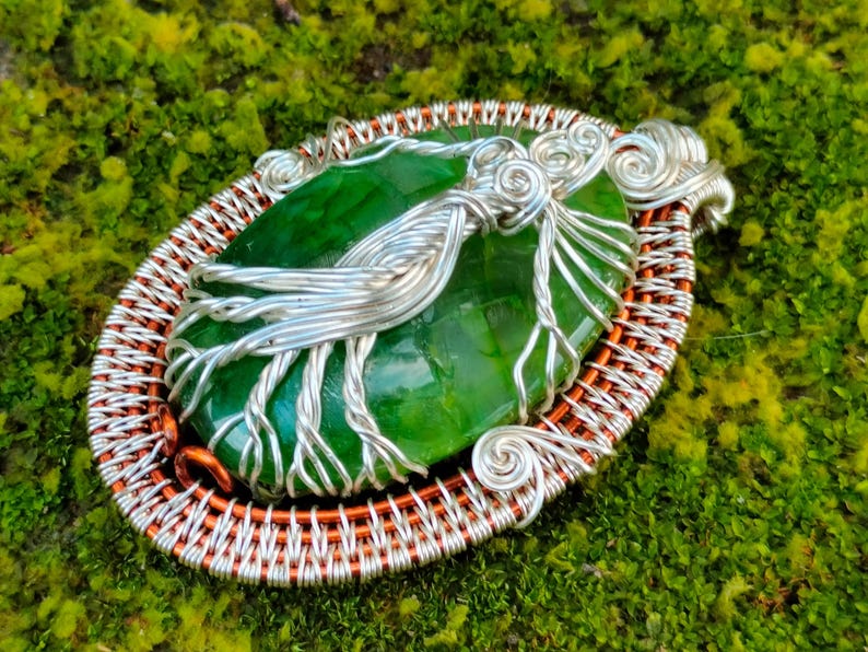 May include: A green gemstone pendant with silver wire wrapped around it, set in a copper wire frame. The pendant is oval shaped and has a smooth, polished surface.