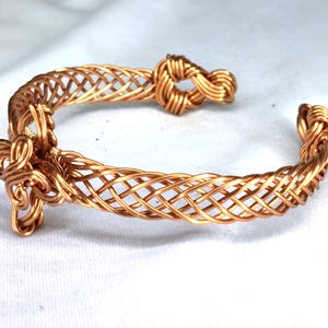 May include: A copper wire cuff bracelet featuring a woven lattice pattern throughout the band. The bracelet ends are finished with intricate, coiled wire knots, creating a continuous, open-ended design made from polished metallic copper wire.
