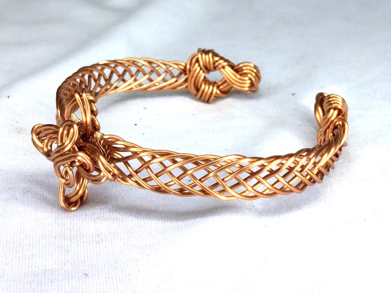 May include: A copper wire cuff bracelet featuring a woven lattice pattern throughout the band. The bracelet ends are finished with intricate, coiled wire knots, creating a continuous, open-ended design made from polished metallic copper wire.