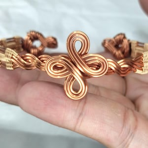 May include: A copper wire cuff bracelet featuring a central four-loop knot design. The band is constructed from twisted copper wire with woven metallic accents along the sides, creating a continuous pattern that wraps around the wrist.
