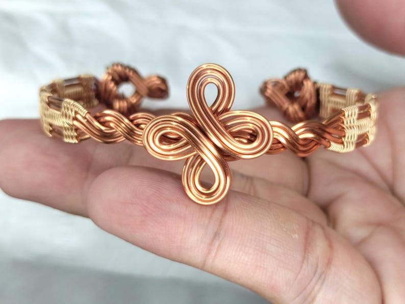 May include: A copper wire cuff bracelet featuring a central four-loop knot design. The band is constructed from twisted copper wire with woven metallic accents along the sides, creating a continuous pattern that wraps around the wrist.
