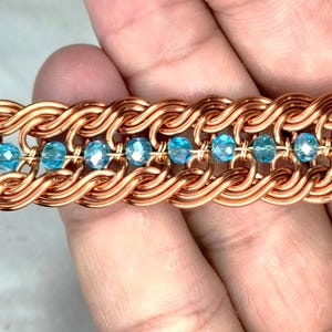 May include: Close-up of a copper-colored bracelet with a woven design. The bracelet features a series of interconnected loops, with small, light blue crystal beads interspersed throughout. The bracelet is a piece of jewelry.