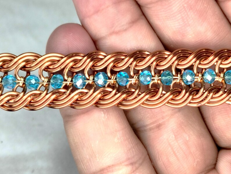 May include: Close-up of a copper-colored bracelet with a woven design. The bracelet features a series of interconnected loops, with small, light blue crystal beads interspersed throughout. The bracelet is a piece of jewelry.