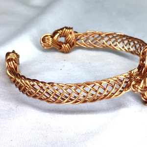 May include: A copper wire cuff bracelet featuring a woven lattice pattern throughout the band. The bracelet has a knot-like design at one end and a more complex, multi-layered wire knot detail at the other end, creating an open-ended structure.