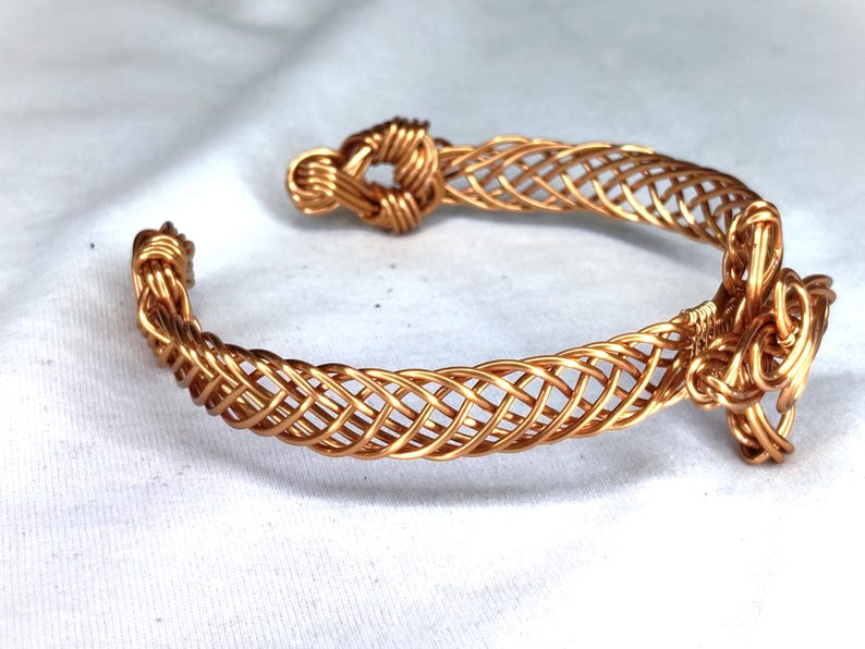 May include: A copper wire cuff bracelet featuring a woven lattice pattern throughout the band. The bracelet has a knot-like design at one end and a more complex, multi-layered wire knot detail at the other end, creating an open-ended structure.