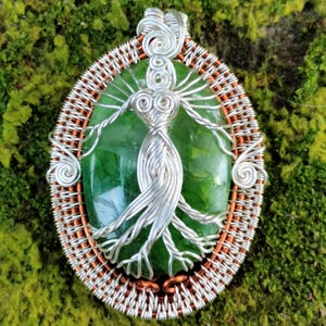 May include: A silver wire wrapped pendant featuring a green gemstone with a tree of life design. The pendant is surrounded by a copper wire frame.