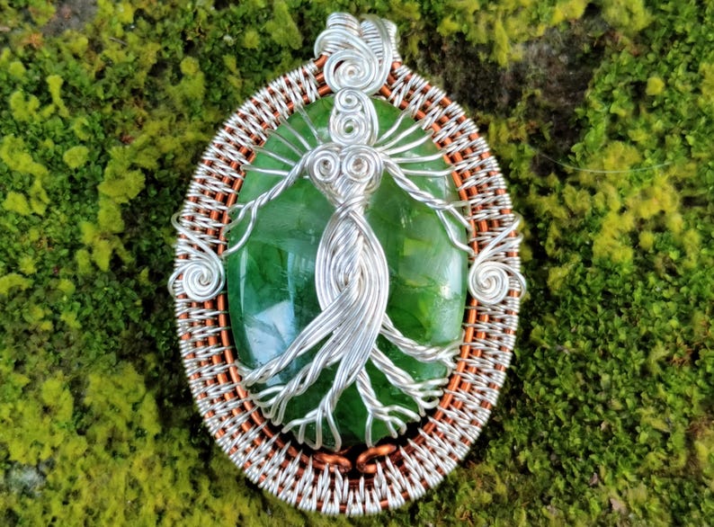 May include: A silver wire wrapped pendant featuring a green gemstone with a tree of life design. The pendant is surrounded by a copper wire frame.
