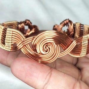 May include: A wire-wrapped cuff bracelet crafted from copper and gold-toned metal wire. The design features a central spiral motif flanked by intricate woven patterns that extend along the band, creating a continuous metallic structure.