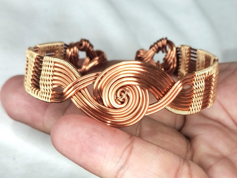 May include: A wire-wrapped cuff bracelet crafted from copper and gold-toned metal wire. The design features a central spiral motif flanked by intricate woven patterns that extend along the band, creating a continuous metallic structure.