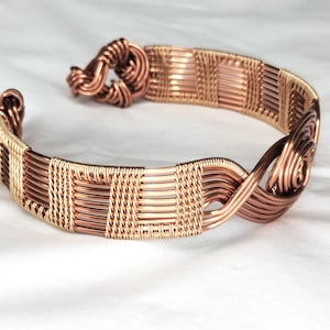 May include: A copper wire cuff bracelet featuring a woven pattern of horizontal and vertical strands. The design includes a coiled knot closure on one end and a spiral detail on the other, showcasing a metallic finish with varying copper tones.