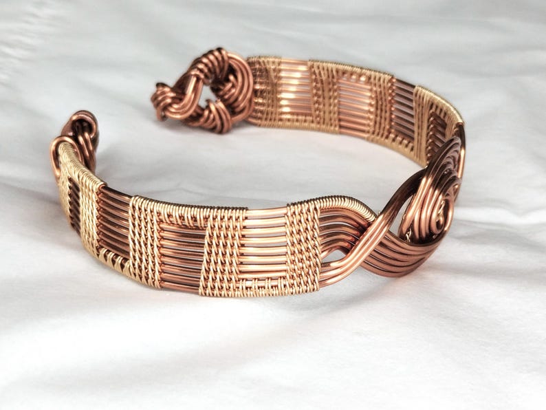May include: A copper wire cuff bracelet featuring a woven pattern of horizontal and vertical strands. The design includes a coiled knot closure on one end and a spiral detail on the other, showcasing a metallic finish with varying copper tones.