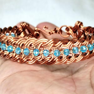 May include: A copper-colored bracelet with a woven design, featuring rows of light blue crystal beads. The bracelet is held in a hand, showcasing its intricate craftsmanship and the contrast between the metal and the beads.