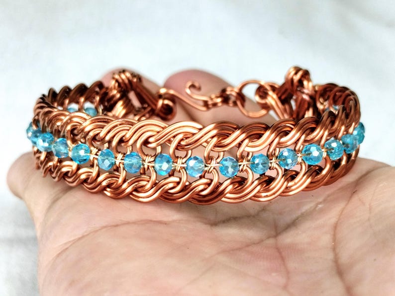 May include: A copper-colored bracelet with a woven design, featuring rows of light blue crystal beads. The bracelet is held in a hand, showcasing its intricate craftsmanship and the contrast between the metal and the beads.