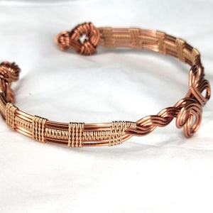 May include: A copper wire cuff bracelet featuring intricate woven patterns and coiled ends. The design consists of multiple parallel copper wires secured by tightly wrapped wire segments, culminating in decorative loops at both open ends.