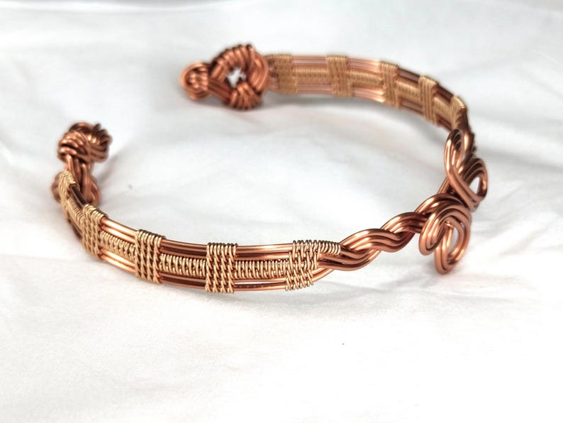May include: A copper wire cuff bracelet featuring intricate woven patterns and coiled ends. The design consists of multiple parallel copper wires secured by tightly wrapped wire segments, culminating in decorative loops at both open ends.