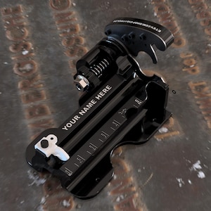 May include: A black metal cigar cutter featuring a spring-loaded mechanism, a white lever, and a built-in ruler marked in inches. The device displays the text 'YOUR NAME HERE' in white lettering and includes a small URL on the top curved edge.