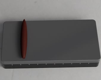 May include: A dark gray rectangular object with a dark red, elongated, conical object resting on top. The rectangular object has a lighter gray border and a series of small, evenly spaced indentations along the bottom edge.