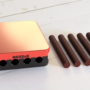 Puede incluir: Una caja de plástico roja y negra con el texto "RG52x6" en la parte delantera. La caja tiene seis agujeros en la parte delantera y está sentada sobre una superficie de madera blanca. Seis clavijas de madera marrón están tumbadas delante de la caja.