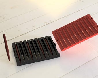 May include: Two red plastic molds for making thin, long, red objects. One mold is open and the other is closed. The closed mold is black and has 12 thin, long, red objects inside.