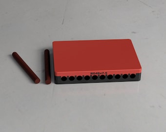 May include: A red rectangular object with a black base and several holes, accompanied by two brown cylindrical rods. The text "RG48x7.5" is printed on the side of the red object. The items are on a light gray surface.