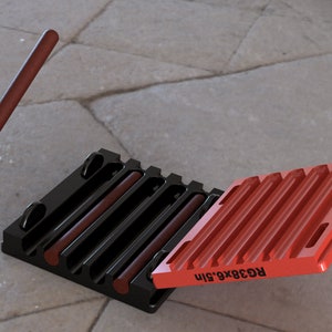 Pode incluir: Um conjunto de calços de roda de borracha pretos e vermelhos. O conjunto inclui dois calços pretos com barras castanhas e um calço vermelho com o texto "RG38x6.5in". Uma barra cilíndrica castanha flutua acima do conjunto.