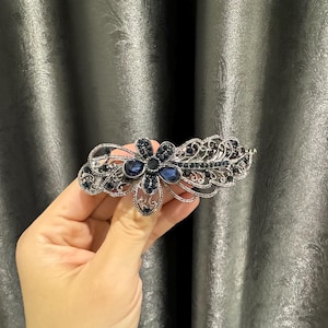 May include: Silver-toned hair clip with a floral design. The clip features a central flower with dark blue gemstones and smaller dark blue accents. The filigree design extends to the sides, creating an elegant accessory.